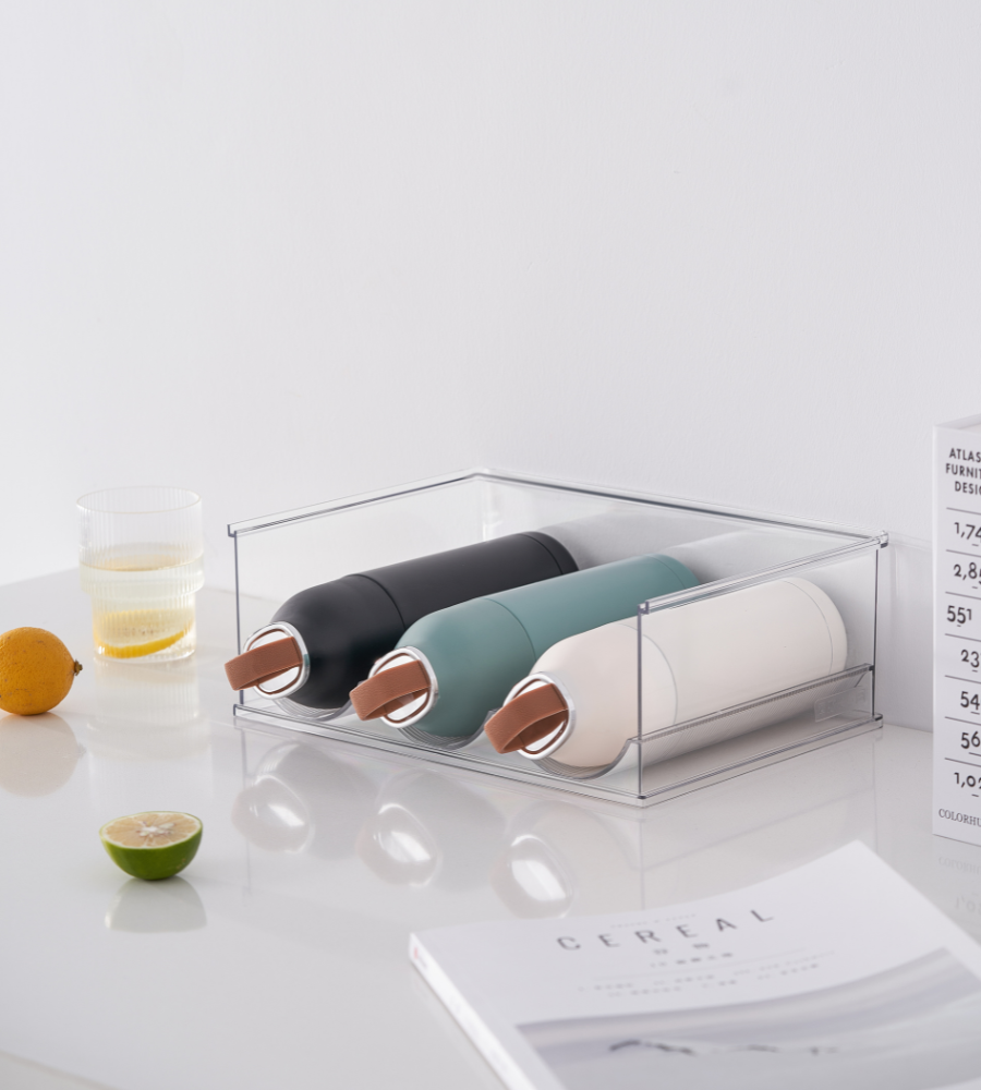 Drei wiederverwendbare Wasserflaschen mit Kupferdeckeln in Schwarz, Grün und Weiß stehen im BNB x VCN Flaschenhalter KLAR - Set 2. Stk auf einer weißen Fläche, neben einem Glas Wasser, einer Zitrone, einer Limette und einer offenen Zeitschrift.