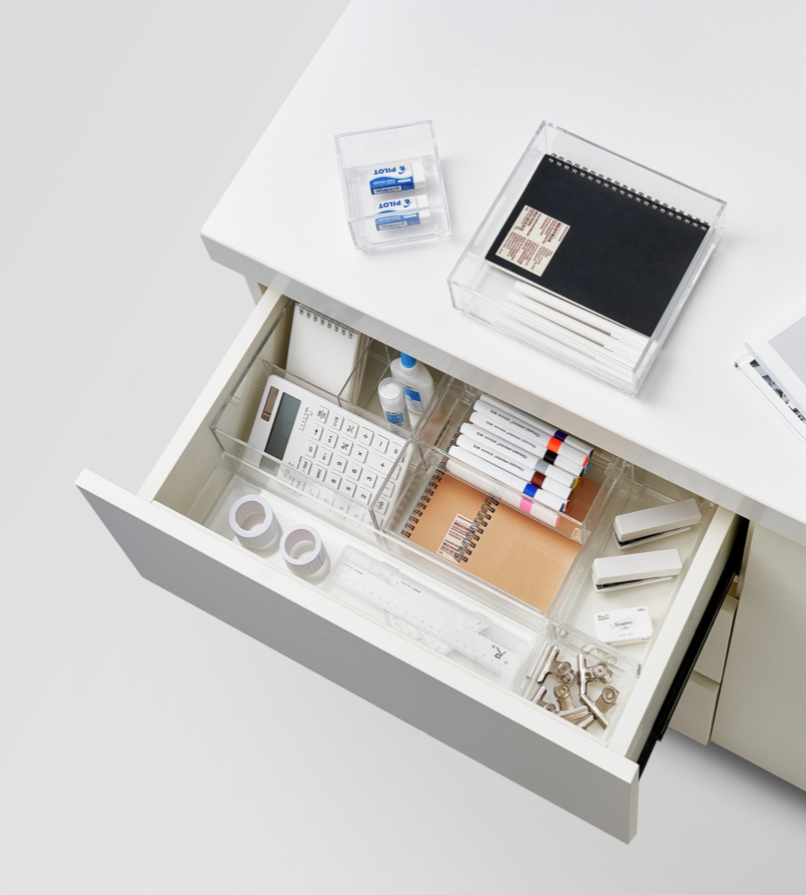 An open white desk drawer is neatly organized with the SLIDE drawer organizer KLAR - HIGH (BNB x VCN, various sizes) and offers space for the most important office utensils such as calculators, pens, highlighters, tape, paper clips, erasers, and small notebooks; other items are stored on the desk above.