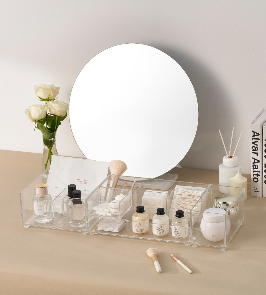 A vanity table with a round mirror, white roses in a vase, skincare and makeup in the BNB x VCN SLIDE drawer organizer CLEAR - HIGH (various sizes), brushes, cotton pads, diffuser sticks, and books on the right-hand side—all on a beige surface.