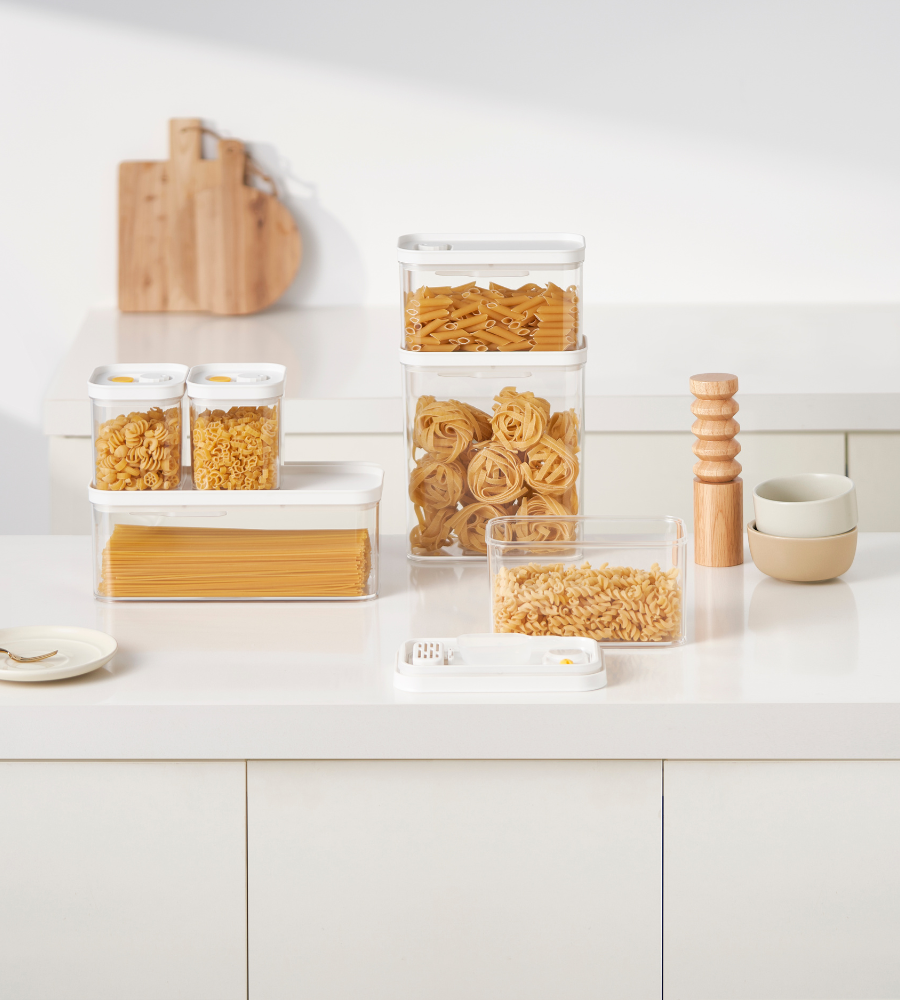 On the kitchen counter are stackable DATEMATE KLAR White storage containers with cooking time timers from BNB x VCN containing pasta, stacked bowls, wooden salt and pepper mills, a plate with a fork, and cutting boards leaning against the wall.