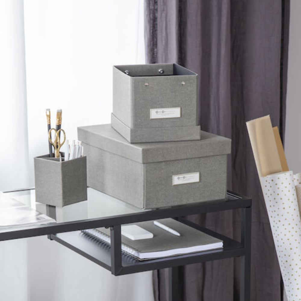 Ein moderner, organisierter Schreibtisch mit der Bigso KARIN Aufbewahrungsbox aus grauem Segeltuch mit Etikettenhalter, einem Stifthalter mit Schere und Bleistiften, einem Notizbuch und aufgerolltem Packpapier für einen minimalistischen Look.