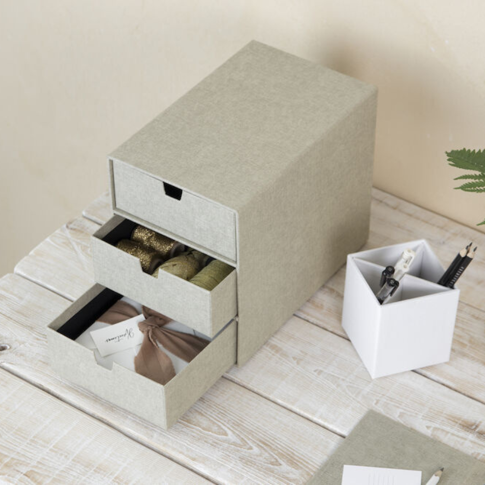 Die graue Bigso INGRID Schubladenbox 3 Fächer - Linnen Canvas steht auf einem weißen Holztisch, in den geöffneten Schubladen liegen Bänder, Stoffe und Geschenkanhänger - perfekt für die Büroorganisation. Daneben steht ein weißer Stiftehalter für Stifte und Scheren.
