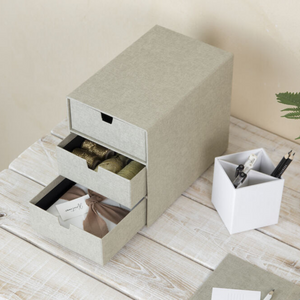 Die graue Bigso INGRID Schubladenbox 3 Fächer - Linnen Canvas steht auf einem weißen Holztisch, in den geöffneten Schubladen liegen Bänder, Stoffe und Geschenkanhänger - perfekt für die Büroorganisation. Daneben steht ein weißer Stiftehalter für Stifte und Scheren.