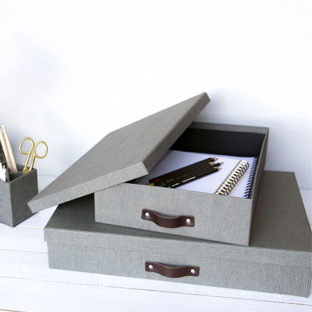 Die Bigso OSKAR Aufbewahrungsbox A4 - Grau Canvas mit braunem Griff ist offen und zeigt Notizbücher und Stifte im Inneren. Daneben hält ein passender Stifthalter Stifte und Scheren auf einer weißen Holzoberfläche organisiert.