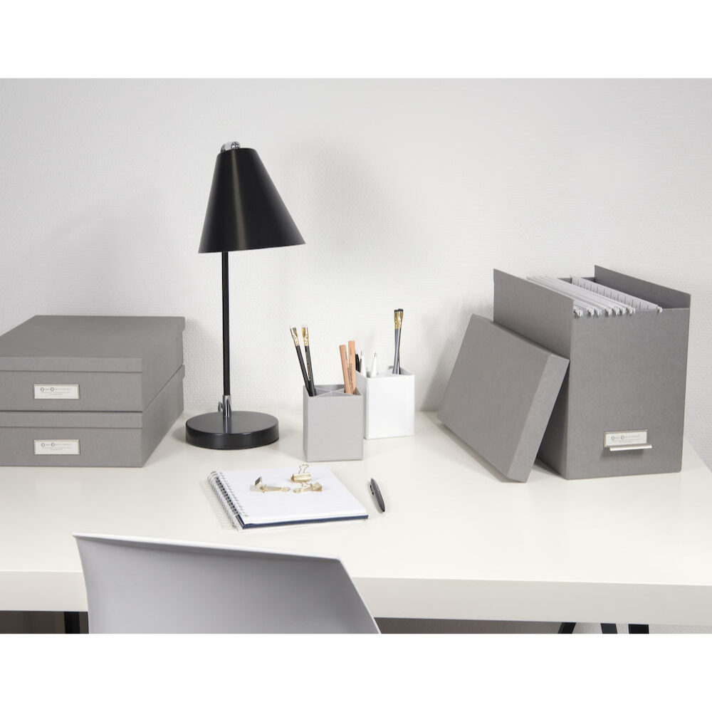 Ein aufgeräumter Bürotisch mit grauen Aufbewahrungsboxen, der Bigso JOHAN Hängeregister-Box - Grau für effiziente Dokumentenorganisation, einem Aktenordner, einer Lampe, zwei Stifthaltern mit Stiften und Bleistiften, einem offenen Notizbuch mit Stift und einem weißen Stuhl davor.