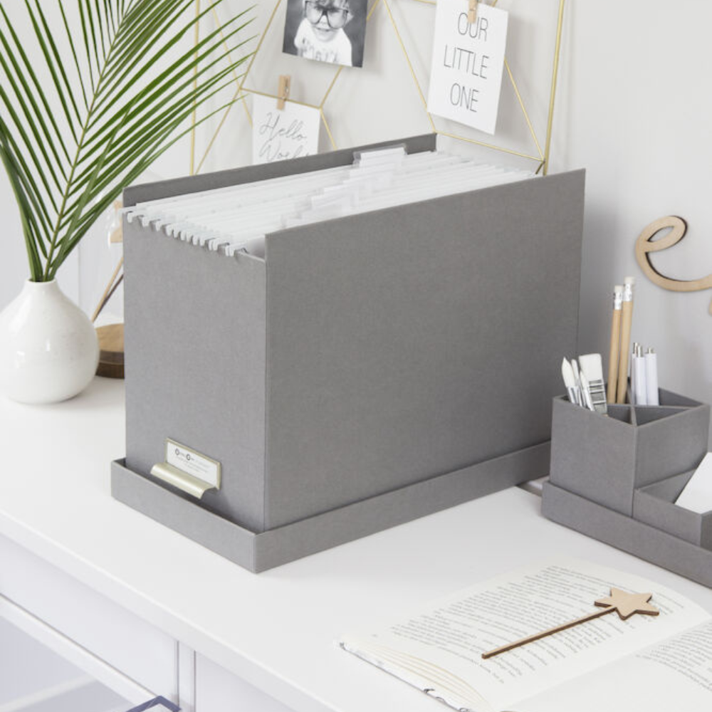 Eine JOHAN Hängeregister-Box in Grau von Bigso mit A4 Hängemappen sitzt auf einem weißen Schreibtisch neben einem passenden Organizer und Stiften; ein aufgeschlagenes Notizbuch, ein sternförmiger Stift und Dekor vervollständigen diesen aufgeräumten Arbeitsbereich.