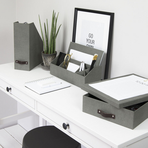 Ein aufgeräumter weißer Schreibtisch mit grauen Büroorganisatoren, einer Bigso OSKAR Aufbewahrungsbox A4 in Grau Canvas, einem Notizbuch, einem gerahmten "GO YOUR OWN WAY"-Zitat, einer Topfpflanze und Schreibwaren, die fein säuberlich angeordnet sind. Darunter steht ein schwarzer Hocker.