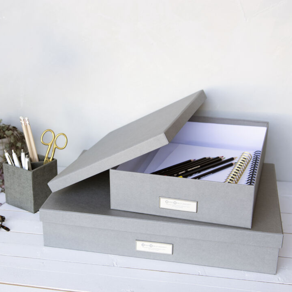 Zwei graue SVERKER Aufbewahrungsboxen A3 von Bigso stehen auf einer weißen Fläche, eine davon offen mit Notizbüchern und Stiften im Inneren. Ein passender Halter daneben hält Scheren, Bleistifte und Stifte - ideal für die stilvolle Aufbewahrung von Dokumenten zu Hause oder im Büro.