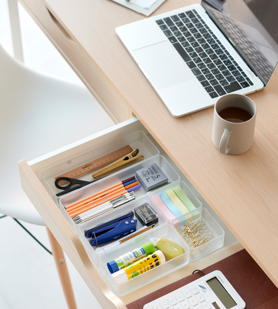 In einer offenen Schublade, die mit dem YAMAZAKI TOWER Schubladen Organizer Weiss L (Set, 2 Stk.) ausgestattet ist, finden Stifte, Schere, Haftnotizen, Klebestifte, Büroklammern und ein Lineal Platz. Auf dem hölzernen Schreibtisch darüber befinden sich ein Laptop, ein Kaffeebecher und ein Taschenrechner.