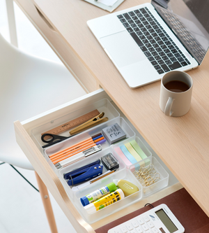 In einer offenen Schublade, die mit dem YAMAZAKI TOWER Schubladen Organizer Weiss L (Set, 2 Stk.) ausgestattet ist, finden Stifte, Schere, Haftnotizen, Klebestifte, Büroklammern und ein Lineal Platz. Auf dem hölzernen Schreibtisch darüber befinden sich ein Laptop, ein Kaffeebecher und ein Taschenrechner.