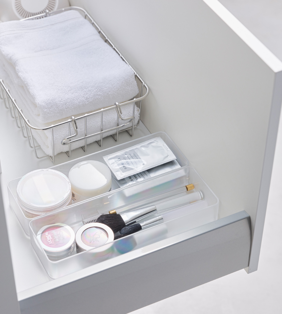 Eine aufgeräumte weiße Schublade mit dem YAMAZAKI TOWER Schubladen Organizer Weiss L (Set 2 Stk.), Metallkorb mit gefalteten Handtüchern und einer durchsichtigen Ablage für Schminkpinsel und Beautyartikel - ideal zum Schubladen organisieren.