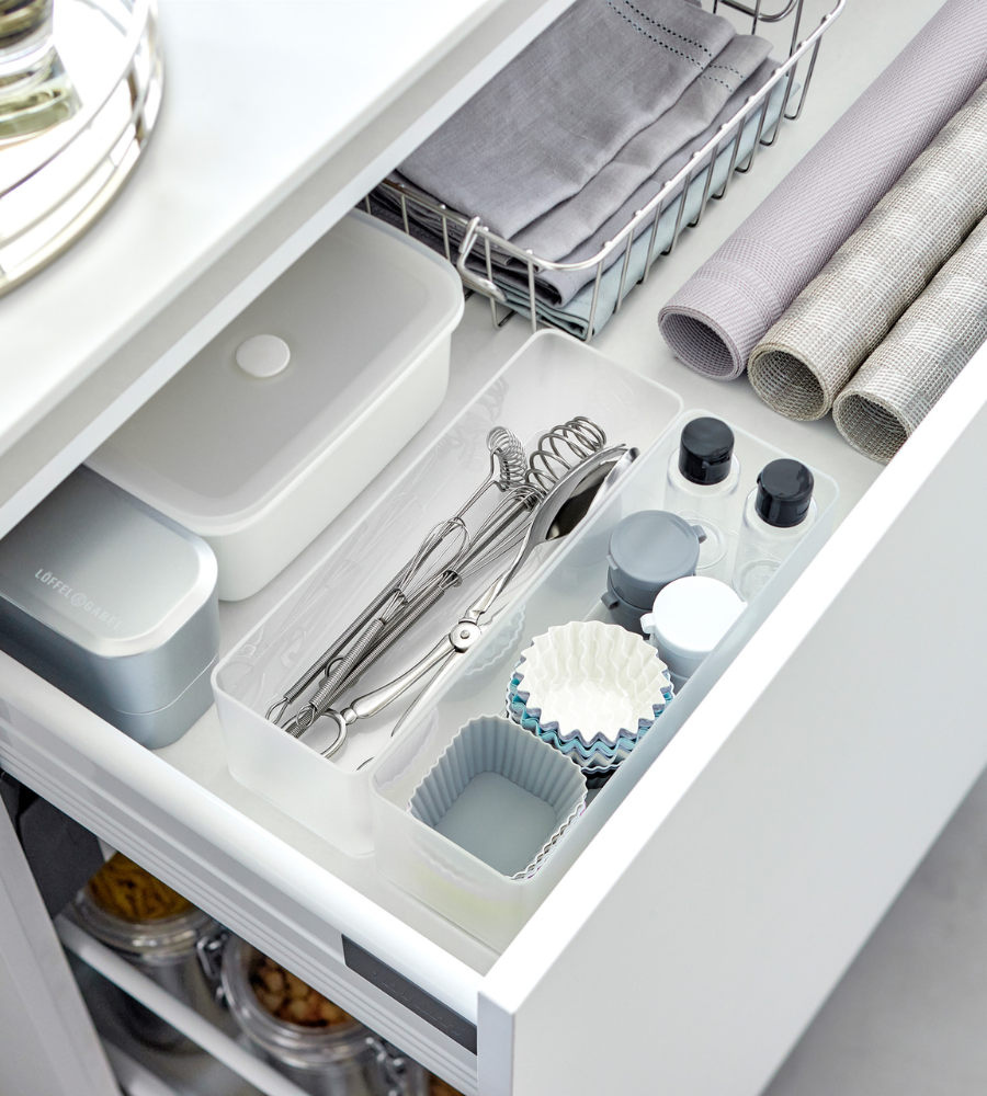 Eine geordnete Küchenschublade mit dem YAMAZAKI TOWER Schubladen Organizer Weiss L DEEP Set (2 Stk.) nimmt Utensilien, Backformen, kleine Flaschen, einen Deckelbehälter, eine Dose und ein Tablett ordentlich auf. Auch gerollte Tischsets und ein Korb mit gefalteten Servietten sind zu sehen.