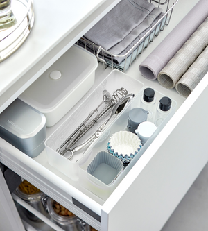Eine geordnete Küchenschublade mit dem YAMAZAKI TOWER Schubladen Organizer Weiss L DEEP Set (2 Stk.) nimmt Utensilien, Backformen, kleine Flaschen, einen Deckelbehälter, eine Dose und ein Tablett ordentlich auf. Auch gerollte Tischsets und ein Korb mit gefalteten Servietten sind zu sehen.