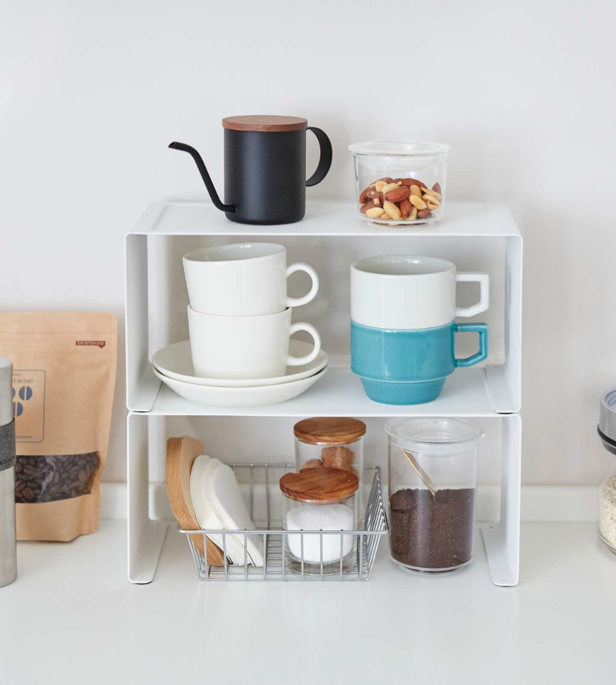 Das TOWER Küchenregal SCHMAL von YAMAZAKI in Weiß organisiert stapelbare Tassen, schwarze Wasserkocher, ein Nussglas, Drahtkorb mit Tellern, zwei Gläser mit Holzdeckeln und Kaffeebehälter - perfekte Raumnutzung in der Küche.