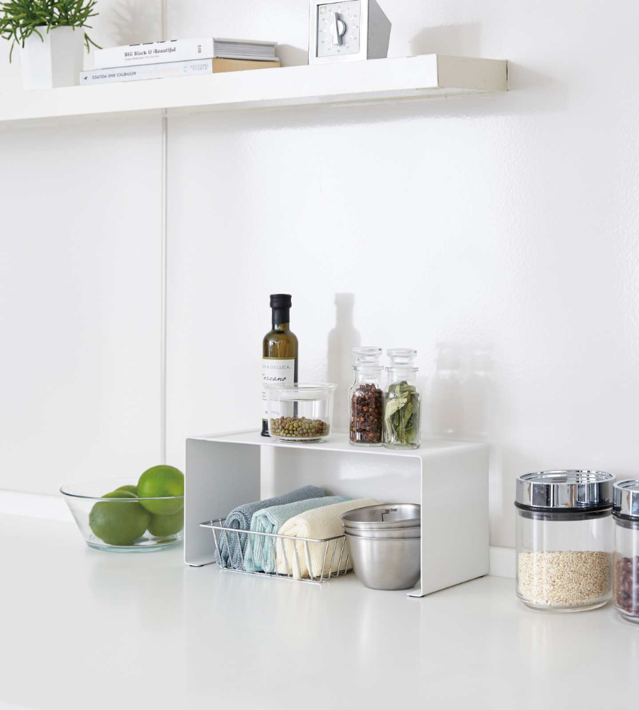 Auf einer modernen weißen Küchentheke steht das YAMAZAKI TOWER Küchenregal SCHMAL - Weiss, das Handtücher, eine Schüssel und Konservendosen aufnimmt. In der Nähe befinden sich grüne Äpfel, Öl, Gewürze und Körner - eine smarte Platznutzung Küche mit Wandregal darüber.
