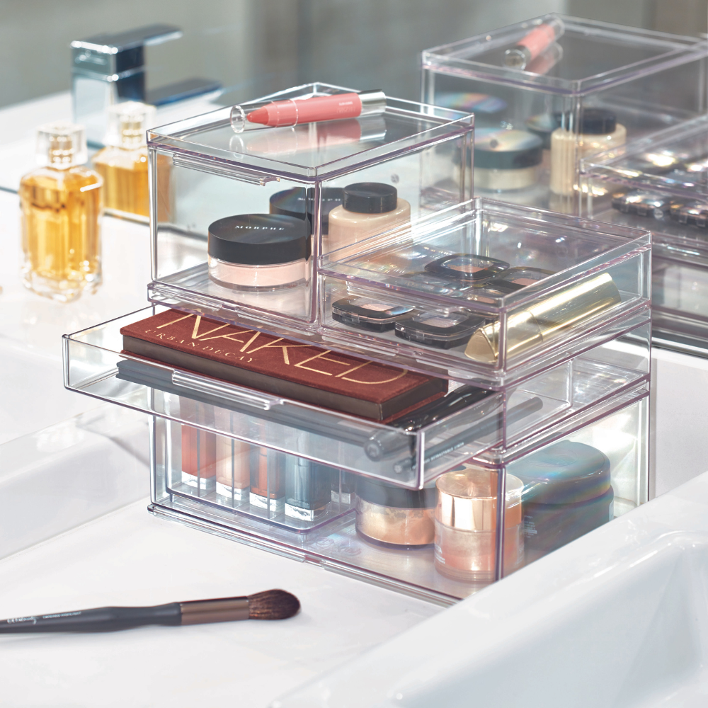 Modularer HOME EDIT Organizer mit Schublade von iDesign, in verschiedenen Größen, gefüllt mit Paletten, Foundation, Lippenstift und Pinseln, stehen neben Parfümflakons und einem Make-up-Pinsel auf dem weißen Bad-Tresen.
