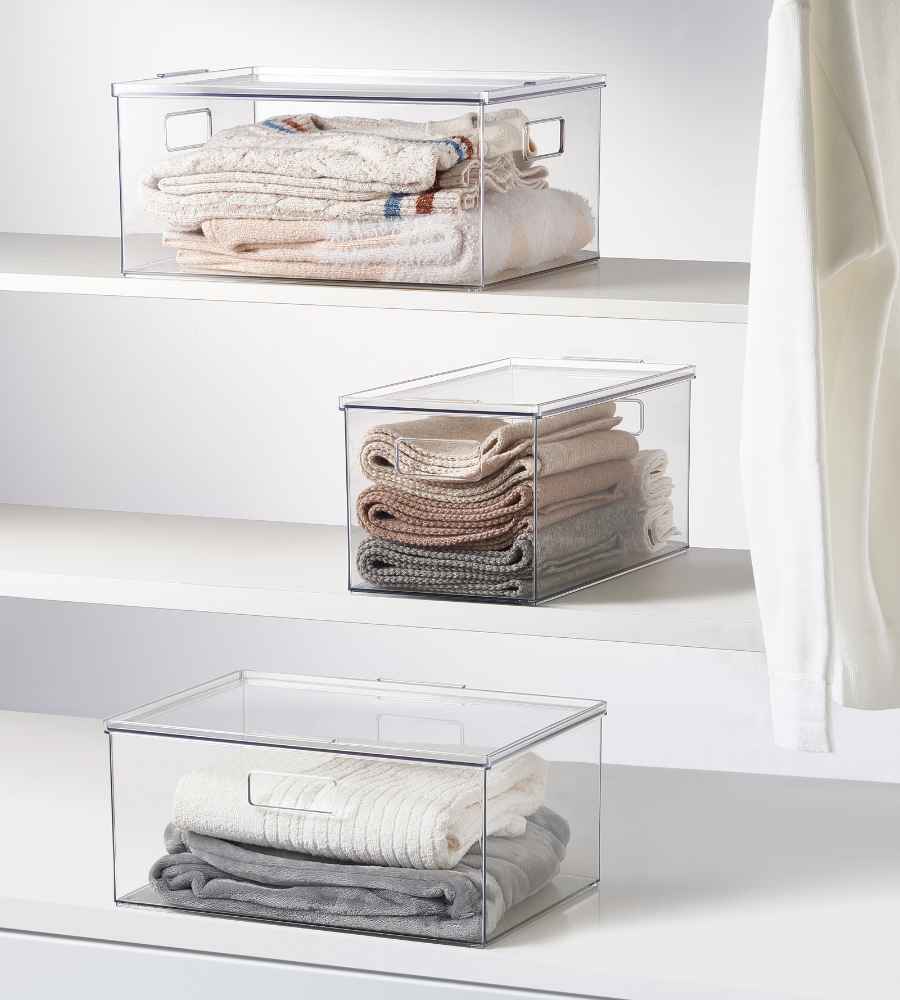 Three stackable storage containers with lids, KLAR by BNB x VCN, in extra-long and various sizes, stand on white shelves filled with clothing; on the right, a white garment hangs over the edge.