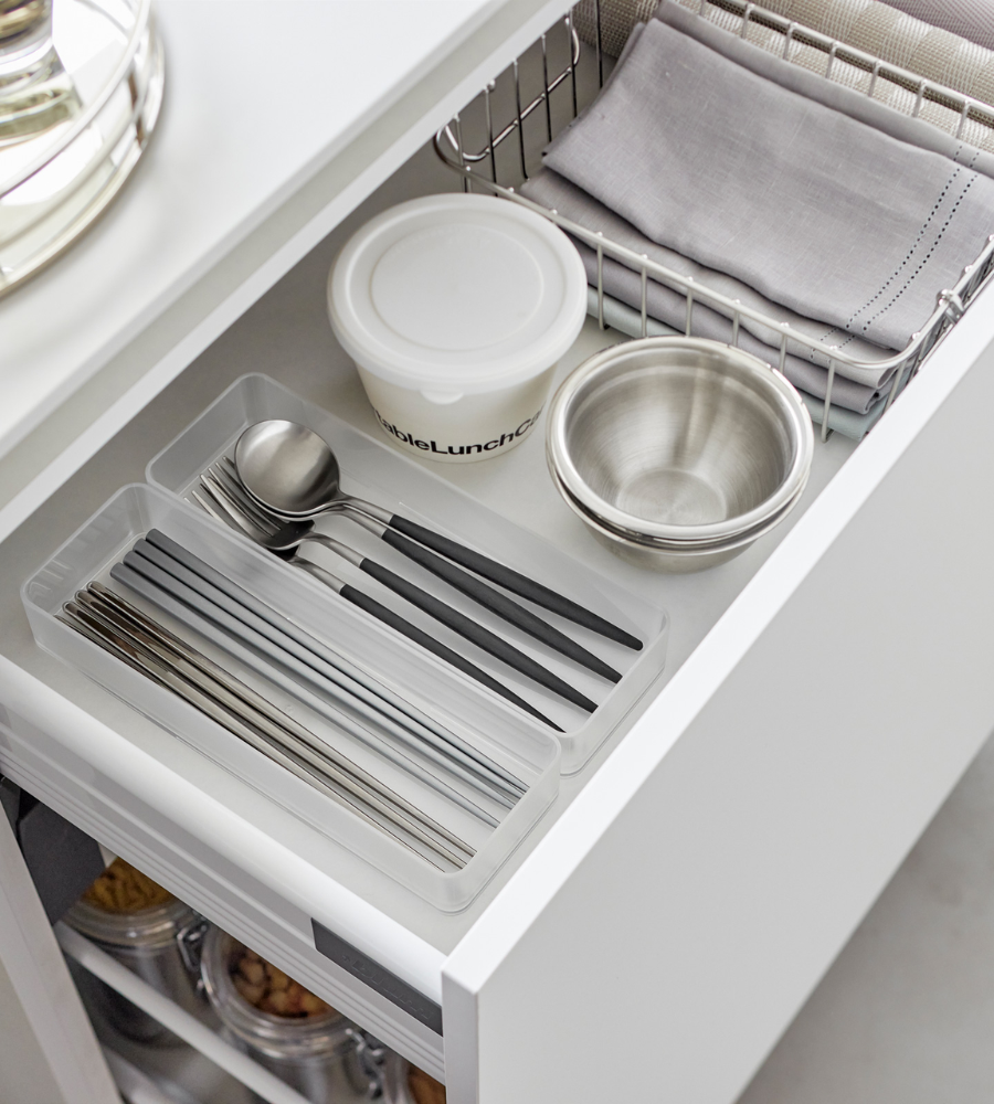 In einer geordneten Küchenschublade sorgt der YAMAZAKI TOWER Schubladen Organizer Weiss L - Set 2 Stk. dafür, dass das Silberbesteck in einem durchsichtigen Tablett neben einem Behälter mit Deckel, einer Metallschüssel und einem Drahtkorb mit gefalteten Stoffservietten oder Tischsets übersichtlich angeordnet ist.