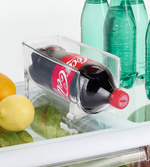 Der BINZ - Flaschenhalter KLAR - Single von iDesign hält zwei Coca-Cola Flaschen liegend auf dem Kühlschrankregal neben grünen Glasflaschen, Orangen, einer Zitrone und Salat.