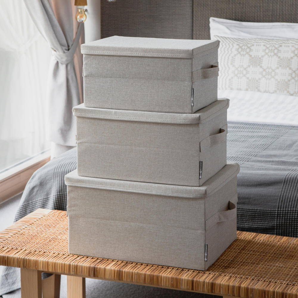 Drei beige SOFT Aufbewahrungsboxen von Bigso (35x26x19cm) sind auf einer geflochtenen Bank in einem Schlafzimmer gestapelt. Diese stilvollen, zusammenklappbaren Stoffaufbewahrungsboxen mit Deckel bieten praktische und elegante Aufbewahrungslösungen.