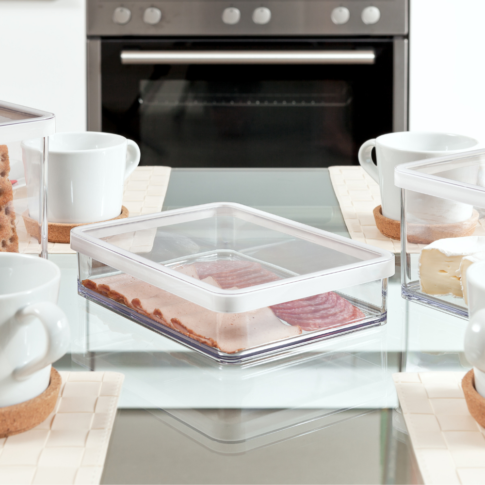 Plastikbehälter mit Wurst, auf Tisch zwischen Tassen, Ofen im Hintergrund.