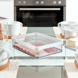 Plastikbehälter mit Wurst, auf Tisch zwischen Tassen, Ofen im Hintergrund.