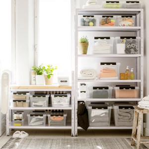 Offene Regale zeigen die Orthex SmartStore Box KLAR 40x30x11cm (7,5L) Flach, BPA-frei und beschriftet, neben gefalteten Handtüchern, Toilettenartikeln und Topfpflanzen in einem hellen Raum mit einem gemusterten Teppich.