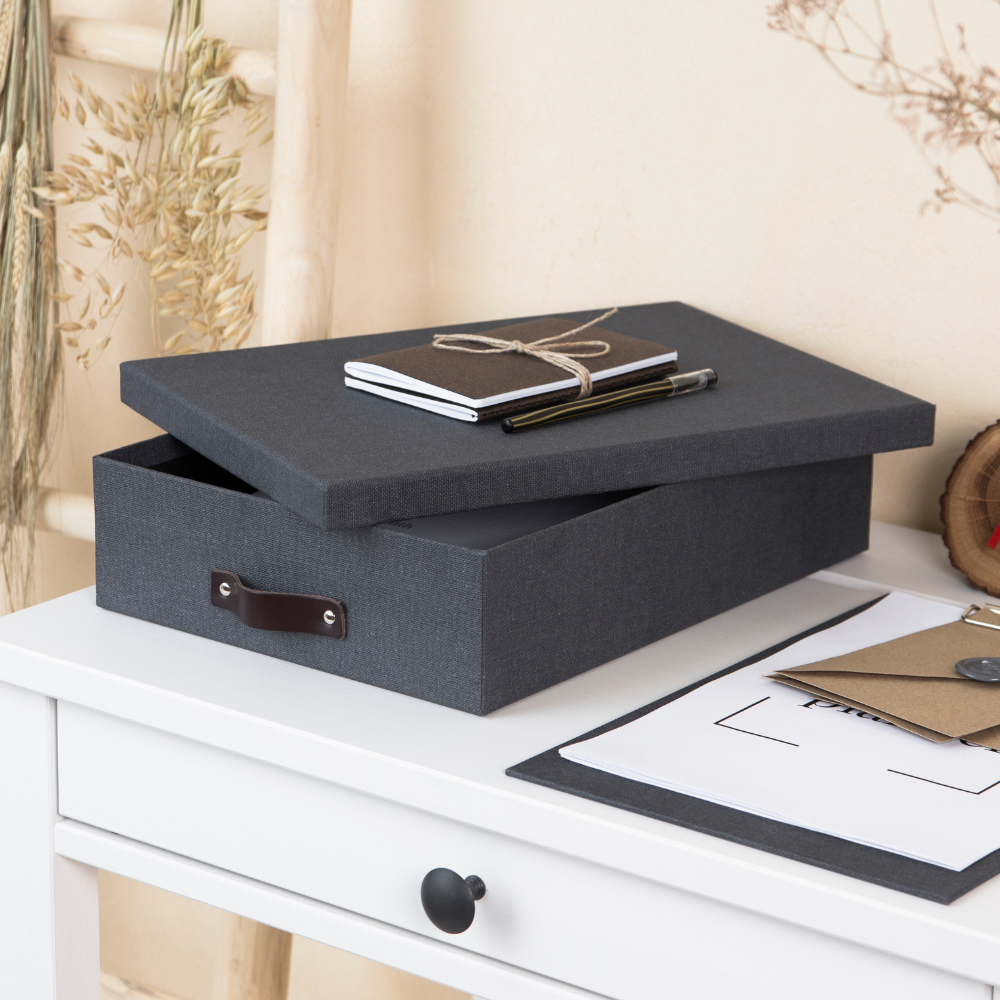Eine Bigso OSKAR Aufbewahrungsbox A4 in Schwarz Canvas mit einem Ledergriff ruht auf einem weißen Schreibtisch. Der Deckel ist leicht geöffnet, darauf liegen ein Notizbuch, ein Stift und ein Bleistift. Papiere, ein Klemmbrett und getrocknete Pflanzen vervollständigen die Szene.