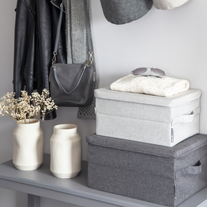 Ein grauer Tisch hält zwei Bigso SOFT Aufbewahrungsbox Beige S (35x26x19cm). Obenauf liegen gefaltete Kleidung und eine Sonnenbrille. Zwei weiße Vasen mit getrockneten Blumen stehen in der Nähe, während eine Lederjacke und eine Handtasche darüber hängen.