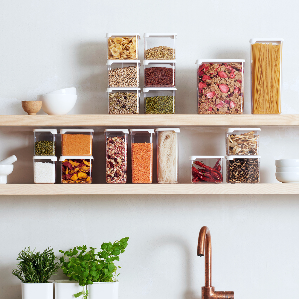 Orthex SmartStore KLAR storage jars in white, filled with grains, pasta, and spices, are neatly arranged on two wooden shelves; herb pots stand below, with a copper faucet underneath.