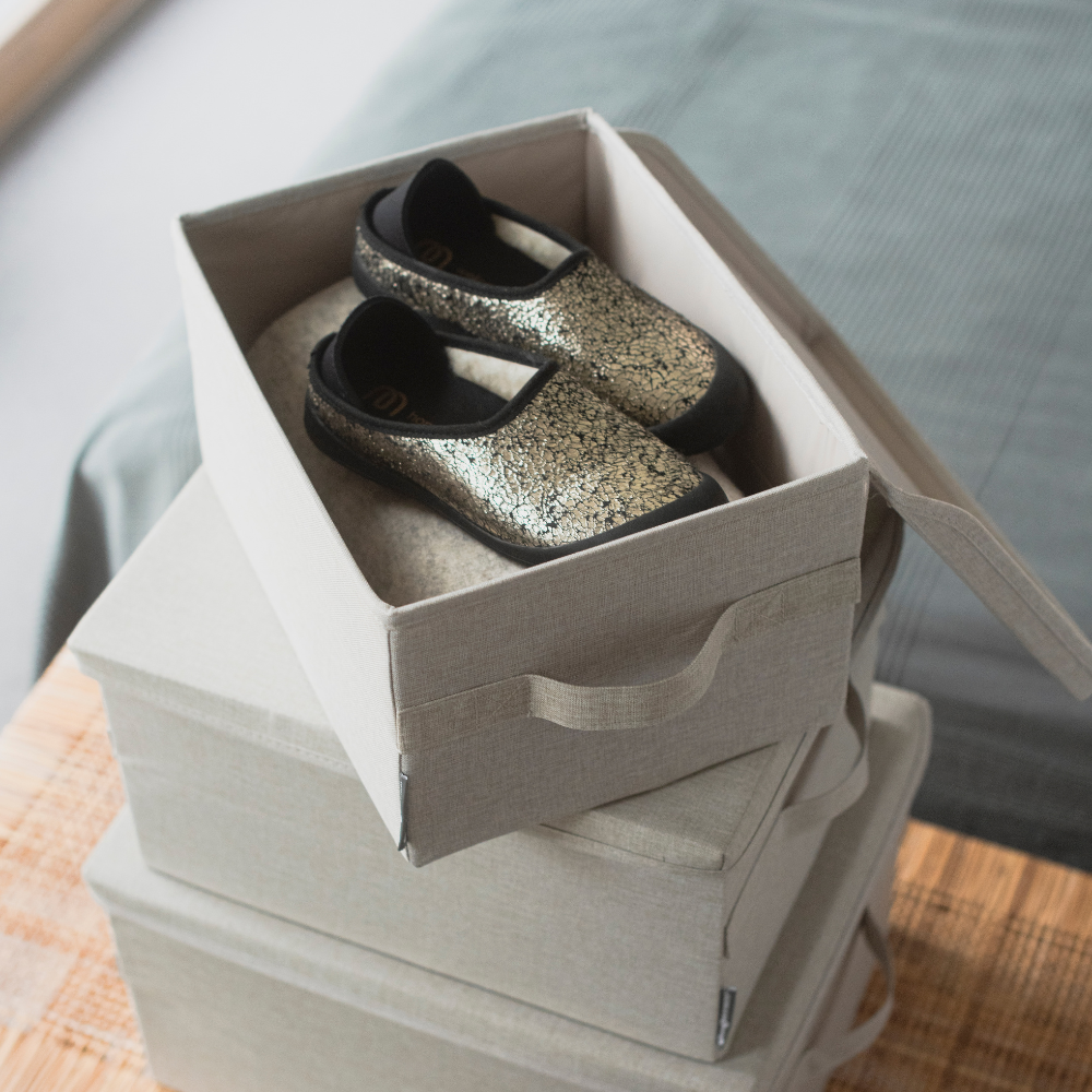 Ein Paar schwarz-gold glitzernde Kinderschuhe sind in einer offenen SOFT Aufbewahrungsbox Beige S (35x26x19cm) von Bigso ausgestellt, gestapelt auf zwei ähnlichen Boxen auf einer hölzernen Oberfläche neben einem Bett.