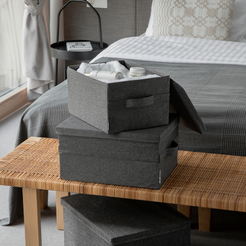 Zwei Bigso SOFT Aufbewahrungsboxen Grau S (35x26x19cm), eine davon offen und mit Toilettenartikeln gefüllt, stehen auf einer geflochtenen Bank am Fuße eines ordentlich gemachten Bettes mit grauer und weißer Bettwäsche in einem modernen Schlafzimmer.